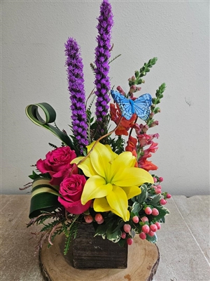 Rustic Springs arrangement with yellow lilies, pink roses, orange snapdragons, purple liatris, and hypericum berries in a cube vase with butterfly accent