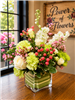 Blush and green garden-style bouquet with roses, carnations, hydrangea, hypericum berries, and stock in a leaf-lined glass cube.
