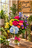 Colorful Wishes Bouquet with blue hydrangea hot pink roses yellow alstroemeria orange carnations blue delphinium purple statice and greenery in a clear glass vase