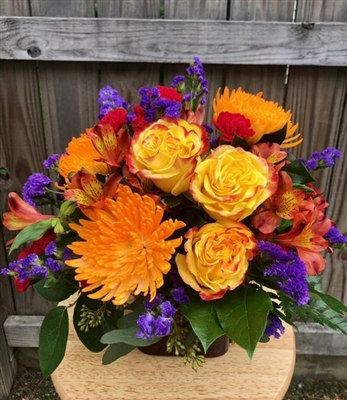 Bright citrus bouquet with yellow roses, red pixie carnations, orange alstroemeria, spider mums, and purple statice arranged in a clear glass cube â€” approx. 13" x 13", all-around.