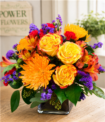 Citrus Sunshine flower bouquet with yellow roses, orange alstroemeria, spider mums, red carnations and purple statice in a glass cube.