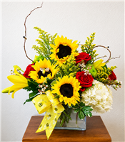 Sunflower and red rose bouquet with yellow lilies, white hydrangea, wax flower, and bee ribbon in a glass cube vase.