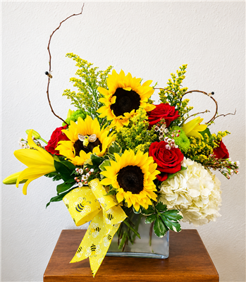 Sunflower and red rose bouquet with yellow lilies, white hydrangea, wax flower, and bee ribbon in a glass cube vase.