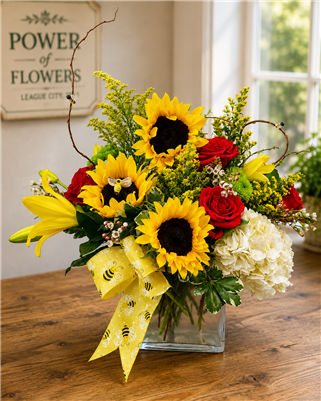Sunflower and red rose bouquet with yellow lilies, white hydrangea, wax flower, and bee ribbon in a glass cube vase.