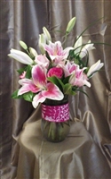 Tall pink bouquet with Stargazer lilies, pink roses, lily grass, and greenery in a clear cylinder vase wrapped with a pink ribbon.