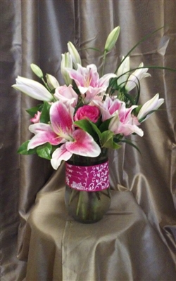 Tall pink bouquet with Stargazer lilies, pink roses, lily grass, and greenery in a clear cylinder vase wrapped with a pink ribbon.