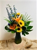 Bright mixed bouquet with sunflowers, orange lilies, blue delphinium, and greenery in a colored glass vase.
