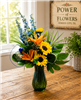 Bright mixed bouquet with sunflowers, orange lilies, blue delphinium, and greenery in a colored glass vase.