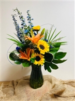 Bright mixed bouquet with sunflowers, orange lilies, blue delphinium, and greenery in a colored glass vase.