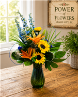 Bright mixed bouquet with sunflowers, orange lilies, blue delphinium, and greenery in a colored glass vase.