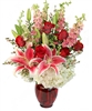 Tall arrangement with Stargazer lilies, red roses, white hydrangea, pink larkspur, and waxflower in a ruby red glass vase.