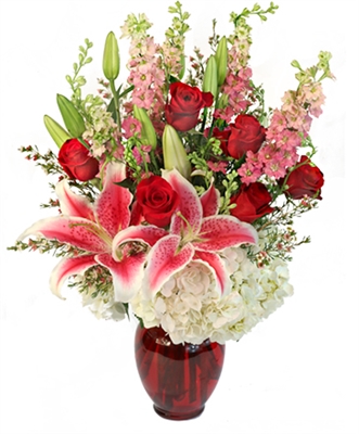 Tall arrangement with Stargazer lilies, red roses, white hydrangea, pink larkspur, and waxflower in a ruby red glass vase.