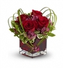 A romantic floral arrangement featuring lush red roses, pink waxflowers, and greenery, artistically designed in a red glass cube vase with decorative bear grass loops.