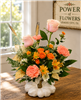 Blush roses, peach carnations, orange spray roses, yellow daisy mums, and limonium arranged in a hand-painted white pumpkin vase with gold stem.