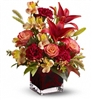 Autumn floral arrangement with orange roses, red Asiatic lilies, burgundy carnations, and seasonal accents in a red glass cube vase