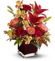 Autumn floral arrangement with orange roses, red Asiatic lilies, burgundy carnations, and seasonal accents in a red glass cube vase