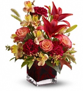 Autumn floral arrangement with orange roses, red Asiatic lilies, burgundy carnations, and seasonal accents in a red glass cube vase