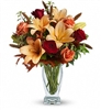 Autumn bouquet with orange lilies, red roses, orange roses, and fall foliage in a clear glass vase.