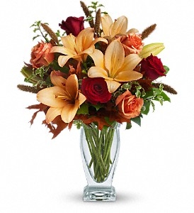 Autumn bouquet with orange lilies, red roses, orange roses, and fall foliage in a clear glass vase.
