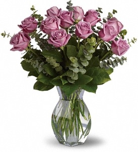 Dozen lavender roses arranged with eucalyptus and greenery in a clear glass vase.