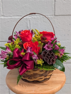Autumn basket arrangement with orange roses, purple alstroemeria, burgundy button mums, solidago, greenery, and a lotus pod with a berry bow.