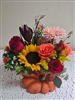 Fall bouquet with sunflowers, red and orange roses, gerbera daisies, and greenery arranged in a ceramic pumpkin vase.