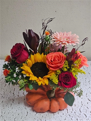 Fall bouquet with sunflowers, red and orange roses, gerbera daisies, and greenery arranged in a ceramic pumpkin vase.