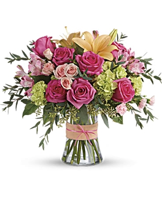 Bright bouquet with hot pink roses, peach lilies, green hydrangea, pink spray roses, alstroemeria, and eucalyptus in a glass gathering vase with a bow.