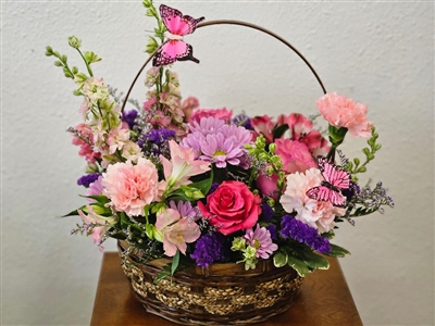 Basket in Blooms arrangement with hot pink spray roses, light pink alstroemeria, snapdragons, mini carnations, waxflower, purple matsumoto asters, Monte Cassino asters, statice, and pittosporum with two butterflies in a wicker basket