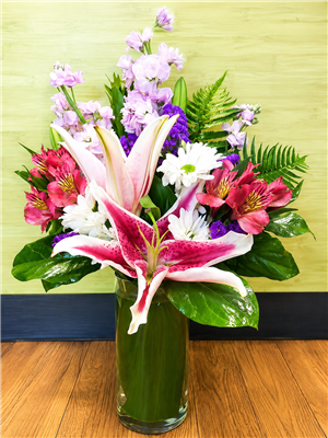 Stargazer lilies with lavender stock, daisies, alstroemeria, and statice in a clear cylinder vase.
